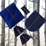 Les sacs en tissu bleu « sport chic » de l’Ensemble, disponibles en plusieurs tailles, se présentent avec art sur un fond à motifs d’arbres noirs et blancs, reflétant l’élégance typique des accessoires d’inspiration lyonnaise.