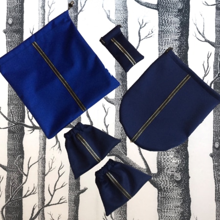 Les sacs en tissu bleu « sport chic » de l’Ensemble, disponibles en plusieurs tailles, se présentent avec art sur un fond à motifs d’arbres noirs et blancs, reflétant l’élégance typique des accessoires d’inspiration lyonnaise.