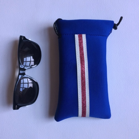 La Mini housse "Sport chic" avec lunettes de soleil noires repose élégamment sur une surface blanche, dotée d'une pochette bleue accentuée d'une rayure rouge et blanche, évoquant les accessoires chics d'un magasin haut de gamme.