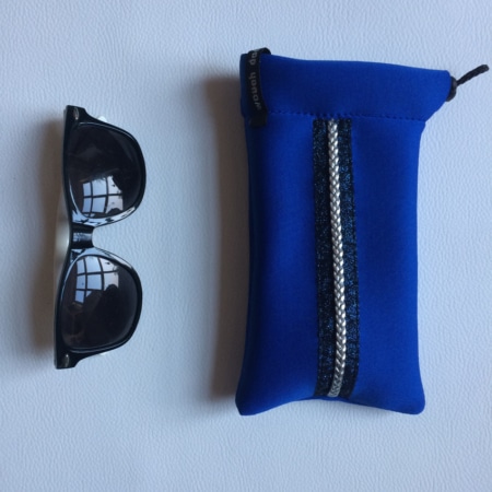 D'élégantes lunettes de soleil noires reposent à côté d'une « Mini housse 'Sport chic' », une pochette en tissu bleu avec une rayure brillante, créant l'accessoire cavalier parfait sur une surface blanche immaculée.