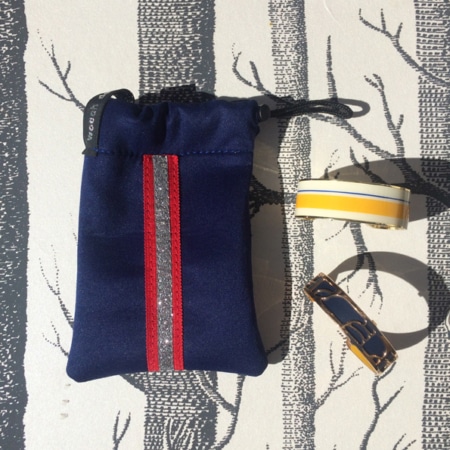 La Mini Housse « Sport chic », un sac à cordon bleu à rayures rouges et argentées, se pose élégamment à côté d'une bande à rayures jaunes et d'un bracelet à motifs dorés, formant ainsi l'ensemble d'accessoires parfait.