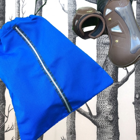 Le sac à cordon bleu et la Housse pour guêtres reposent élégamment sur un fond à motifs d'arbres noir et blanc, évoquant l'esprit d'un cavalier prêt pour une aventure équestre.