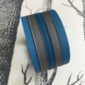 Le « Bracelet cuir manchette » affiche un design rayé bleu et gris reposant élégamment sur un fond texturé, dégageant un charme cavalier.