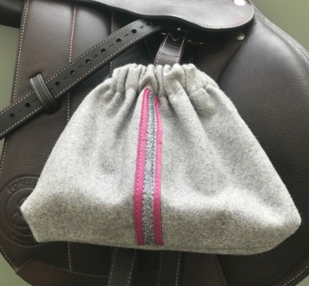 La « Housses d’étriers « sport chic » » est une élégante pochette à cordon grise à rayures roses et argentées, élégamment posée sur une selle en cuir marron foncé, un accessoire indispensable pour tout passionné d’équitation.