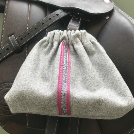 La « Housses d’étriers « sport chic » » est une élégante pochette à cordon grise à rayures roses et argentées, élégamment posée sur une selle en cuir marron foncé, un accessoire indispensable pour tout passionné d’équitation.