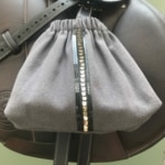 Le « Housses d’étriers « sport chic » » est un sac à cordon gris avec une bande verticale pailletée noire sur des accents de cuir marron aux détails surpiqués, idéal pour le cavalier soucieux du style.