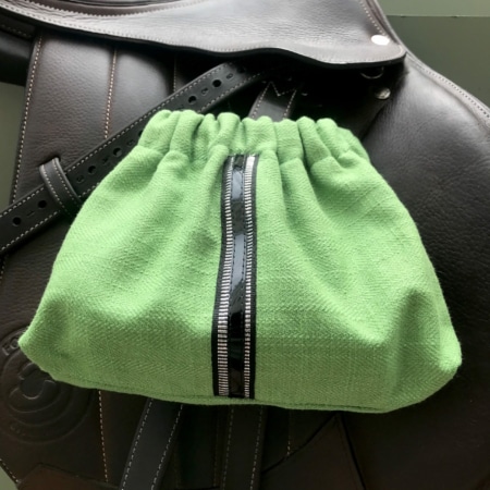 Un sac à cordon Housses d’étriers « sport chic » vert avec fermeture éclair noire repose élégamment sur une selle en cuir noir, parfaitement accessoirisée pour le cavalier.