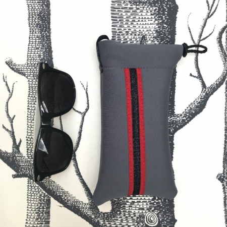 Les Housses d’étriers « Sport » reposent à côté d’une pochette grise à rayures rouges et noires, sur fond d’illustrations d’arbres. Cet accessoire chic ajoute une élégance cavalière à n’importe quelle tenue, alliant style et praticité sans effort.