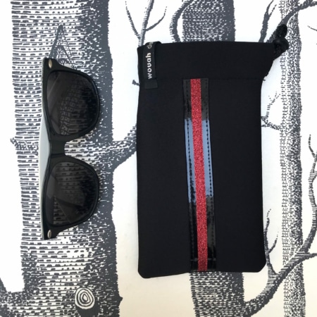 Des lunettes de soleil à côté d'une pochette chic noire « Housses d'étriers 'Sport' » à rayures rouges et bleues, conçue comme un accessoire incontournable pour les cavaliers modernes, sur fond monochrome à motifs d'arbres.